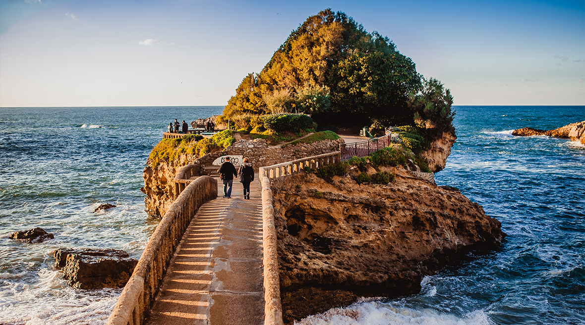 bridge leading over a rocky cove to a rock jutting out into the sea covered with trees and shrubbery at sunset